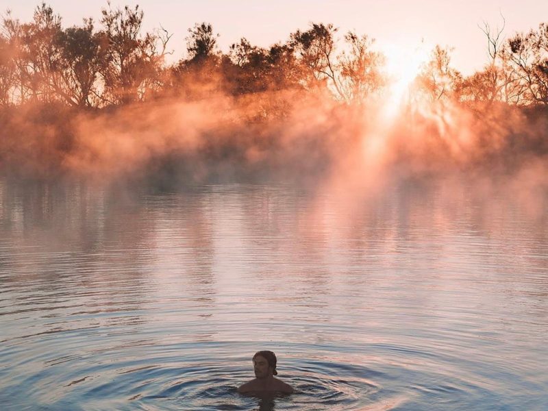 A man swims in Dalhousie Springs, behind him the sun shines through the morning fog.
