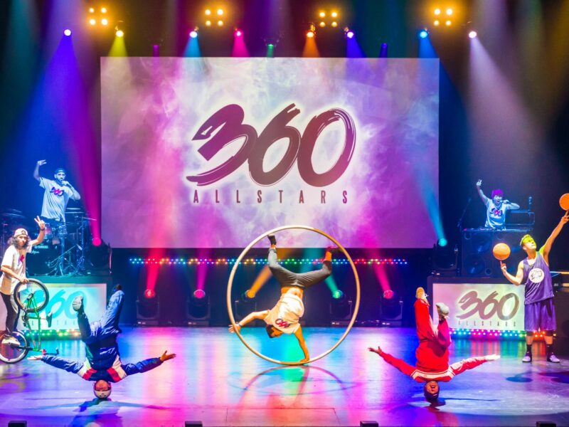 A group of acrobats doing tricks on stage lit by multi coloured lighting.
