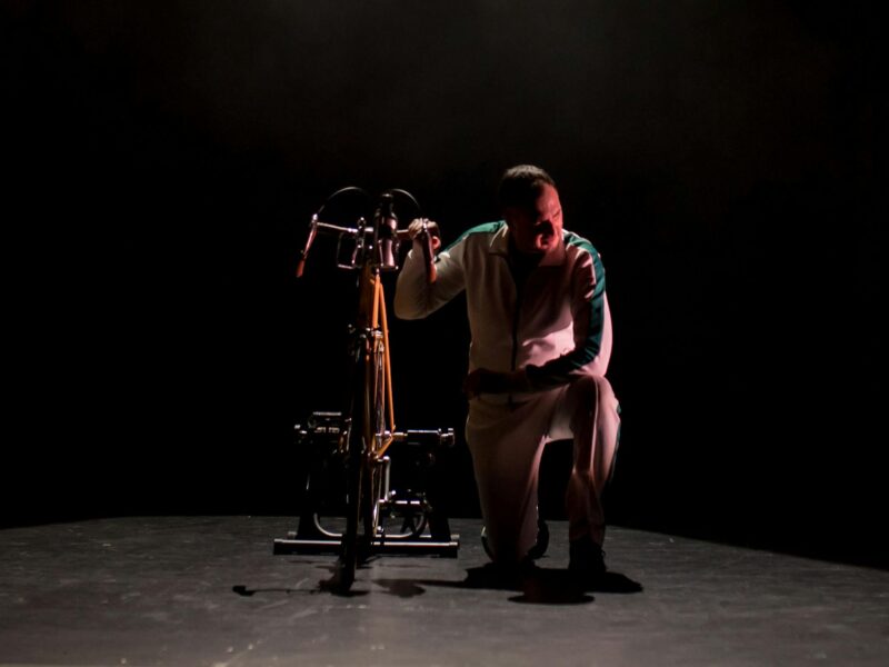 A man in a white tracksuit kneeling next to a bicycle on a black stage under a spotlight