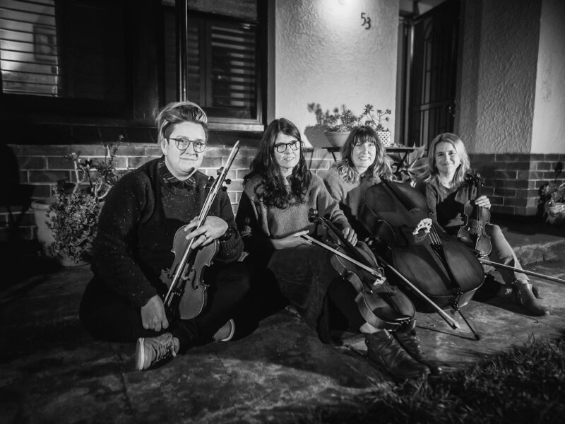 Four people sitting on the ground outside of a house holding various string instruments