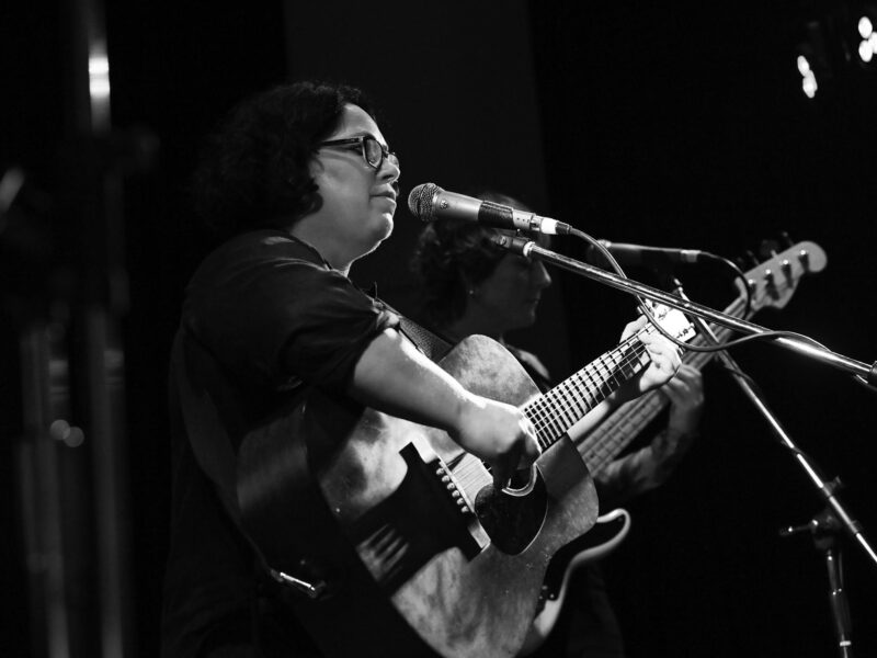 Nancy Bates holding a guitar in front of a microphone, behind her are other musicians.