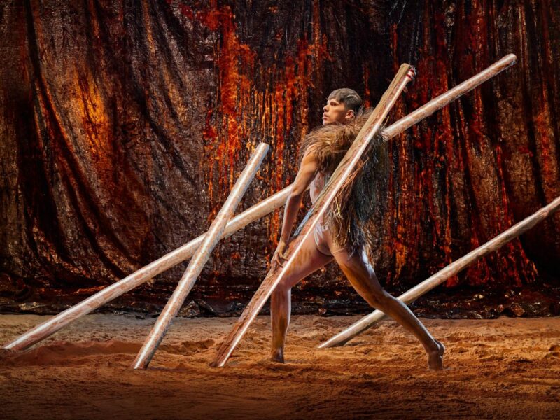 A man weaves through 4 poles protruding from the sandy ground against a metallic backdrop