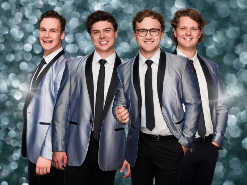 Band members in silver jackets and ties, posing with smiles against a sparkling background.