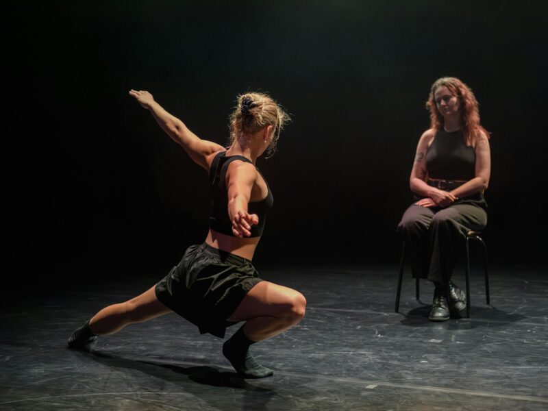 A woman sits in a chair watching a dancer perform for her in the foreground.