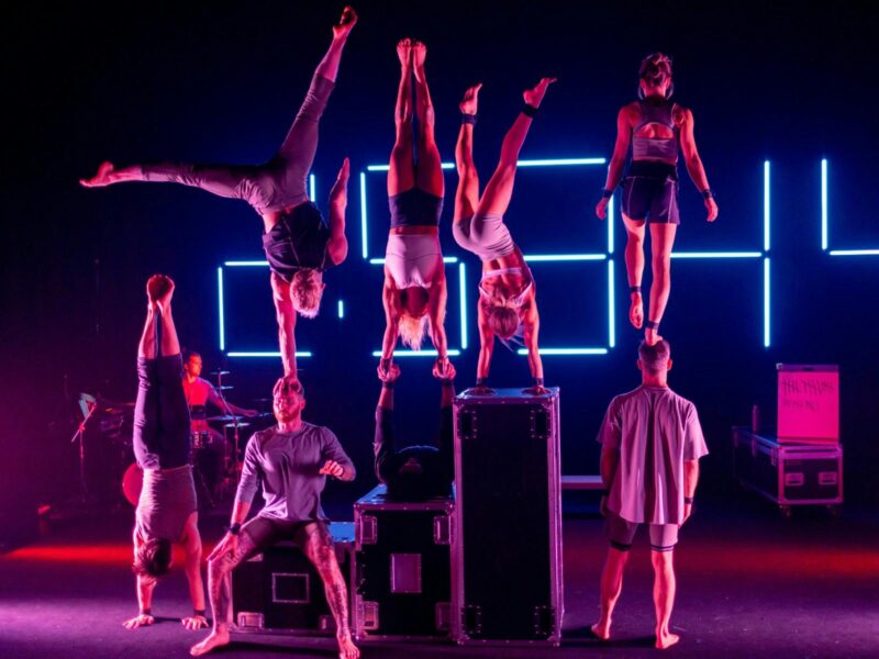 A group of acrobats performing stunts lit by coloured lighting with a neon clock in the background.